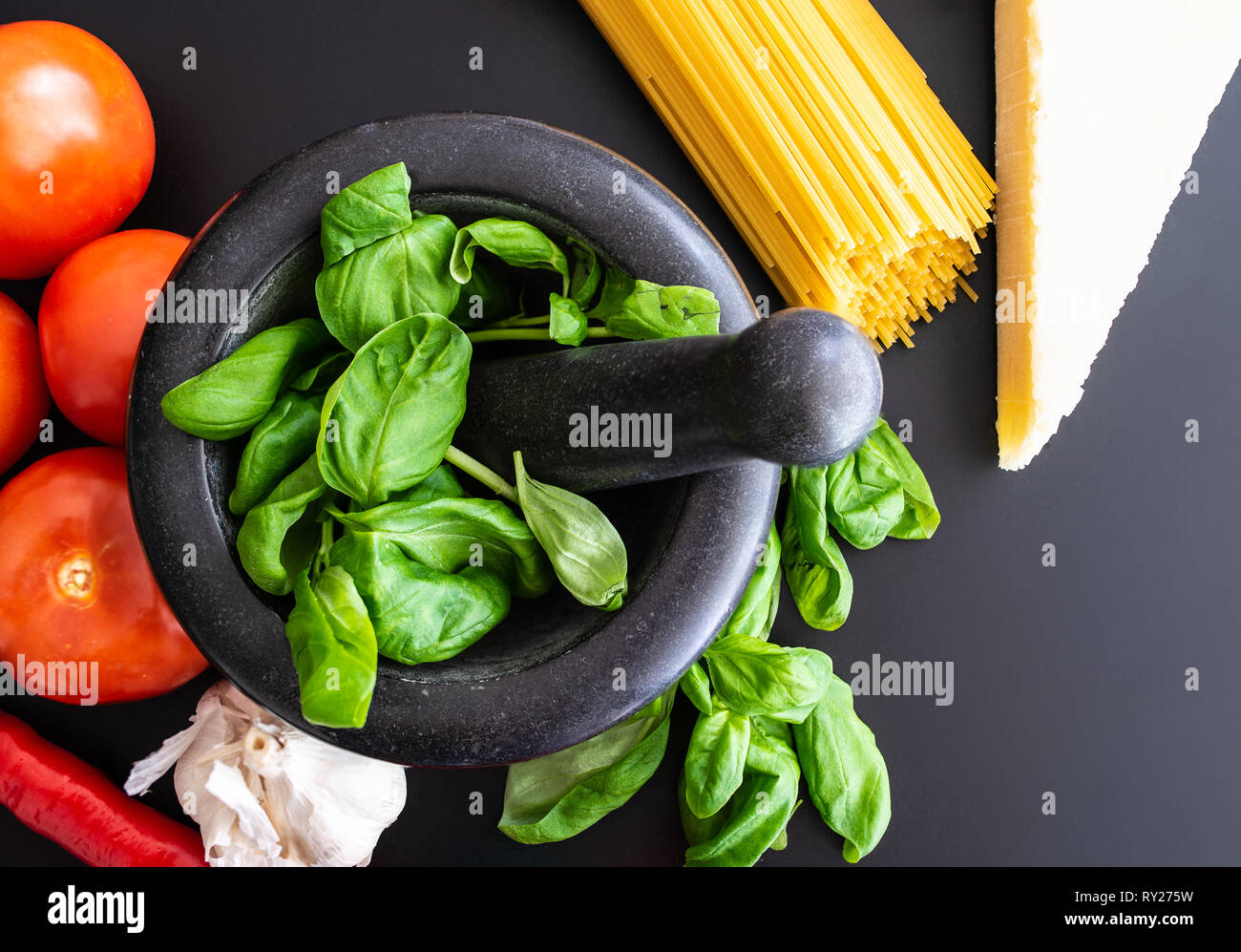 preparing italian pasta with fresh basil in mortar, tomatoes, garlic, pepper and Parmesan on dark kitchen counter Stock Photo