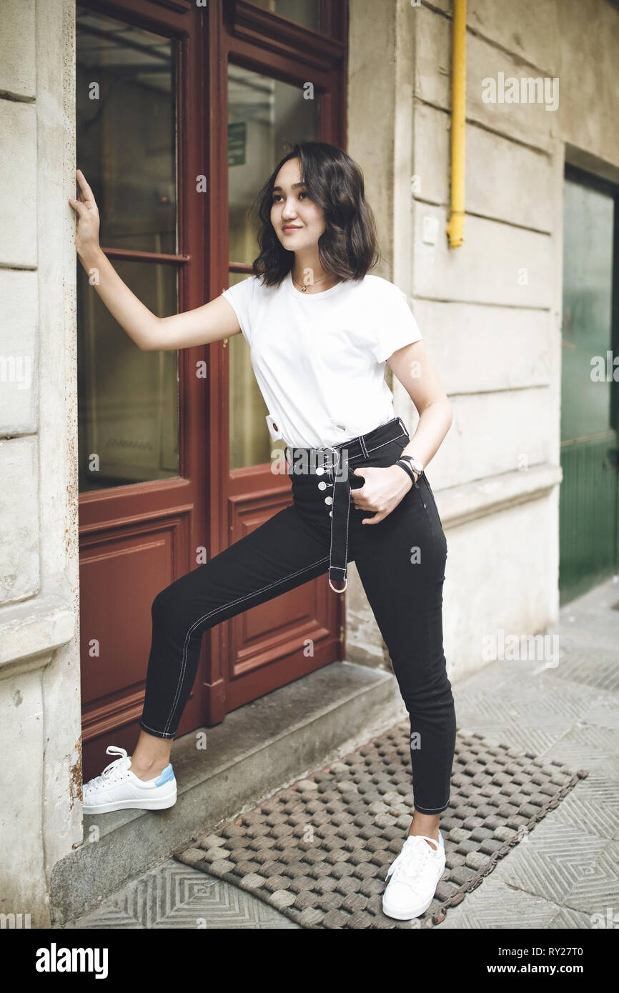 Cheerful optimistic asian girl in a white t-shirt and black jeans, waiting  near closed door at the entrance, smiling. Street portrait Stock Photo -  Alamy