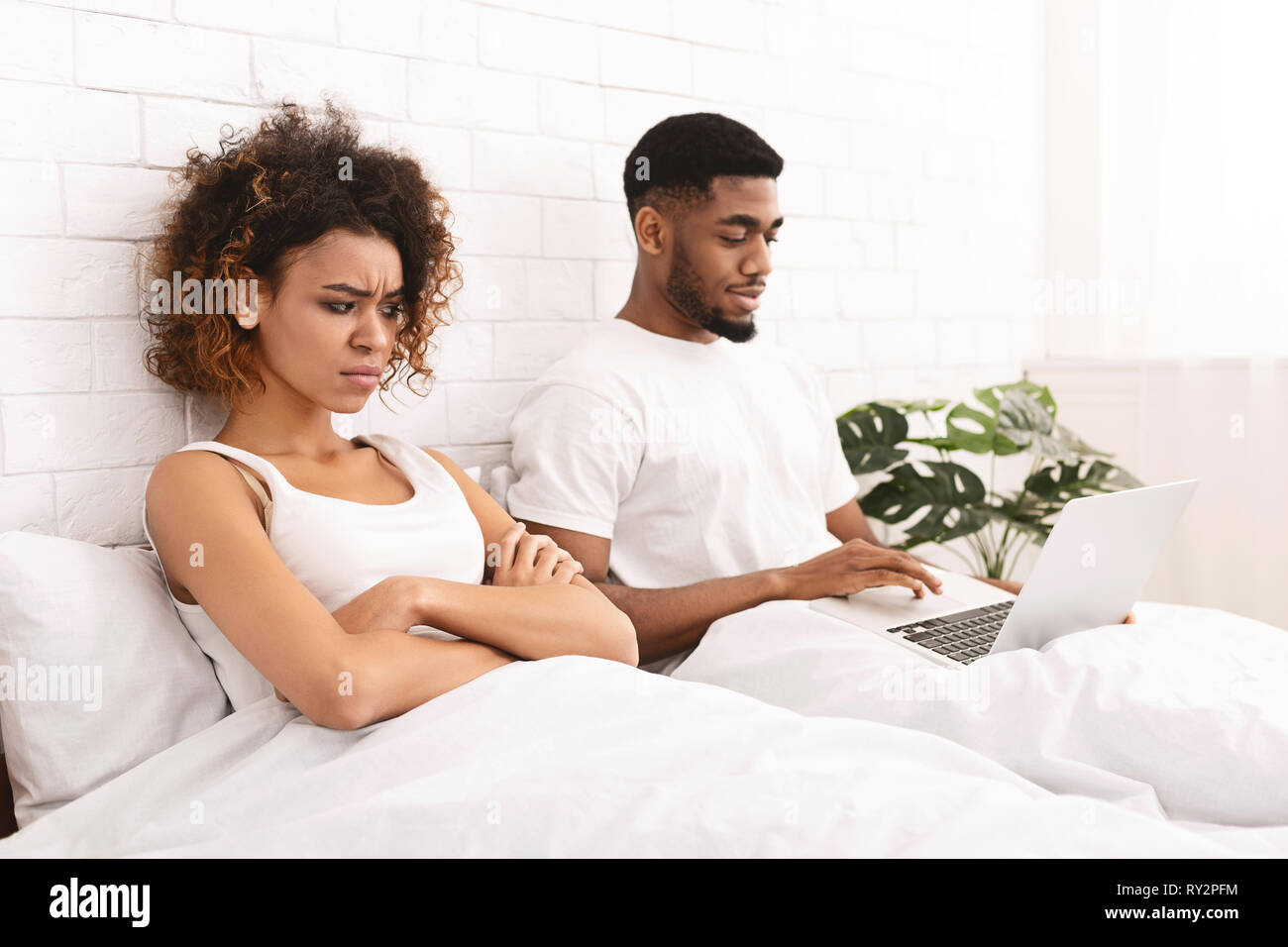 Addicted young man in bed with laptop, woman looking angry Stock Photo