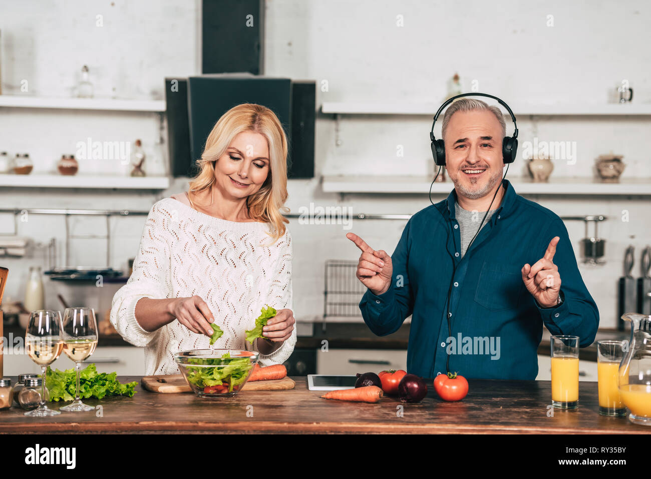 cheerful woman preparing food near happy husband listening music in headphones in kitchen Stock Photo