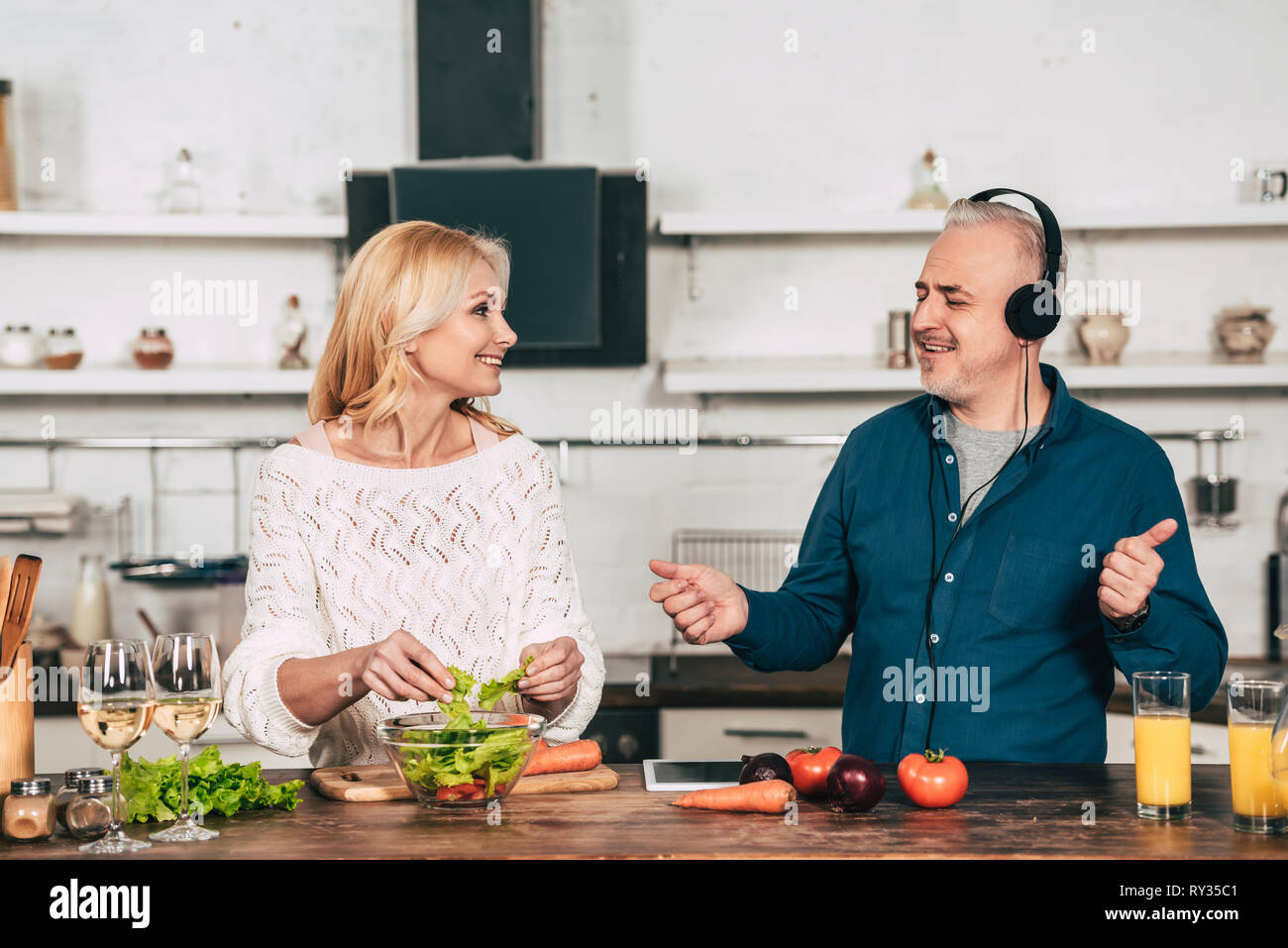 happy woman preparing food near happy husband listening music in headphones in kitchen Stock Photo