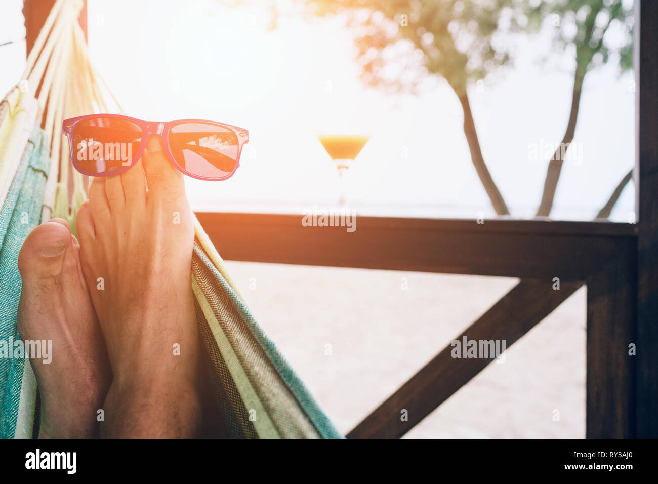 Young man lying in a hammock at a sunny beach by ocean. Sunglasses on the feet. Stock Photo