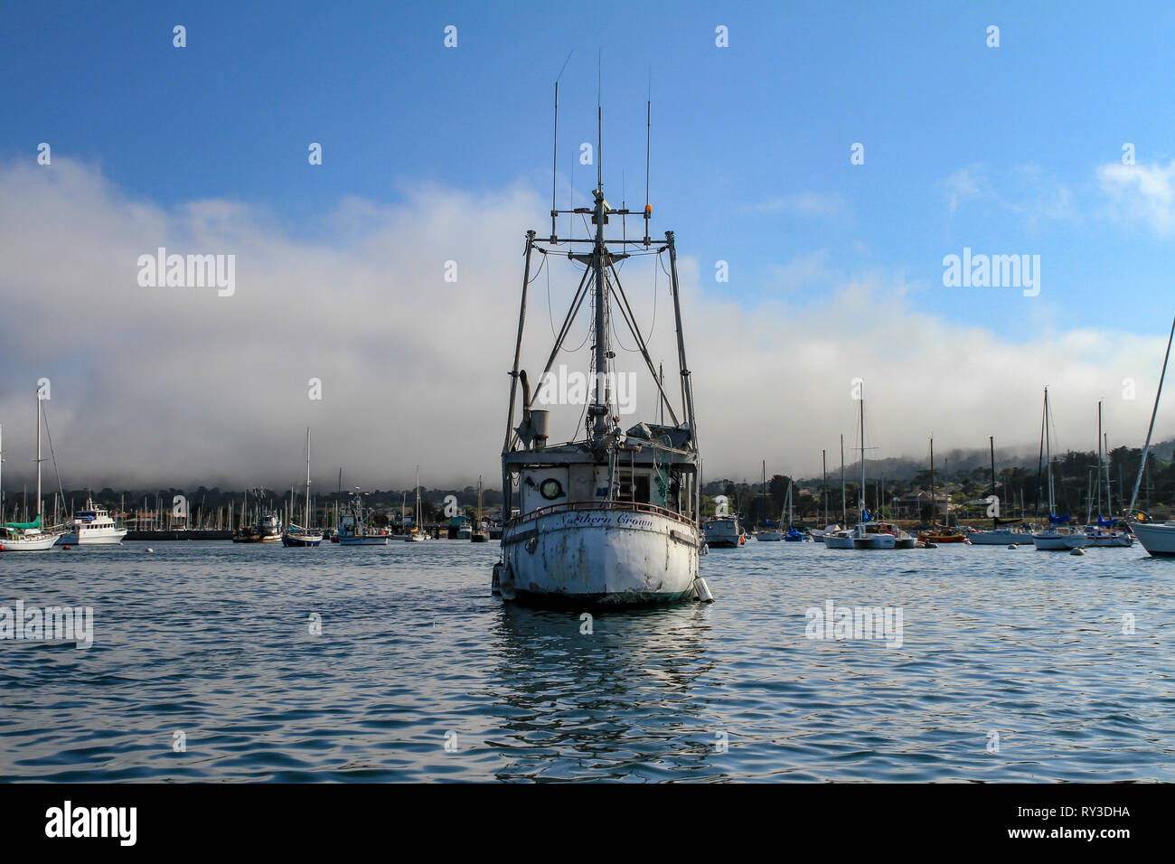 Middle sized boat with radio antennas and old white ship boards floating on calm water in the bay, viewed from its front. Yachts and sailboats moored  Stock Photo