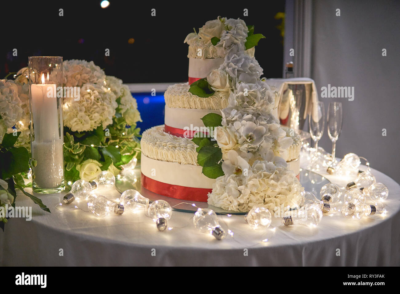 A three-tiers white wedding cake with rose decors on a table decorated with white candles and light bulbs. Landscape format. Stock Photo