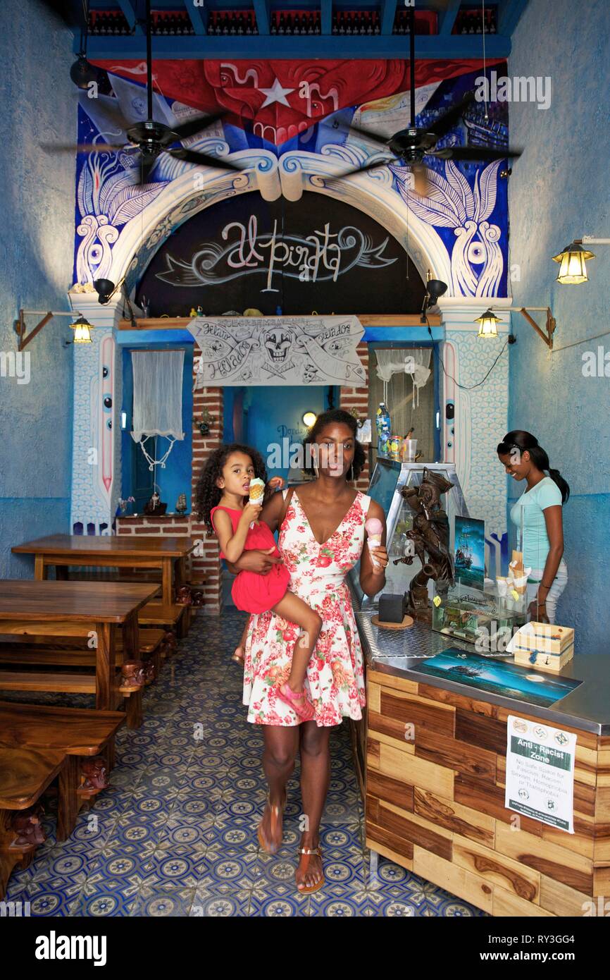 Cuba, La Havana, old Habana, listed as World Heritage by UNESCO, black woman in flower dress with her child in arms and an icecream in hand at the ice-cream maker Al Pirata Stock Photo