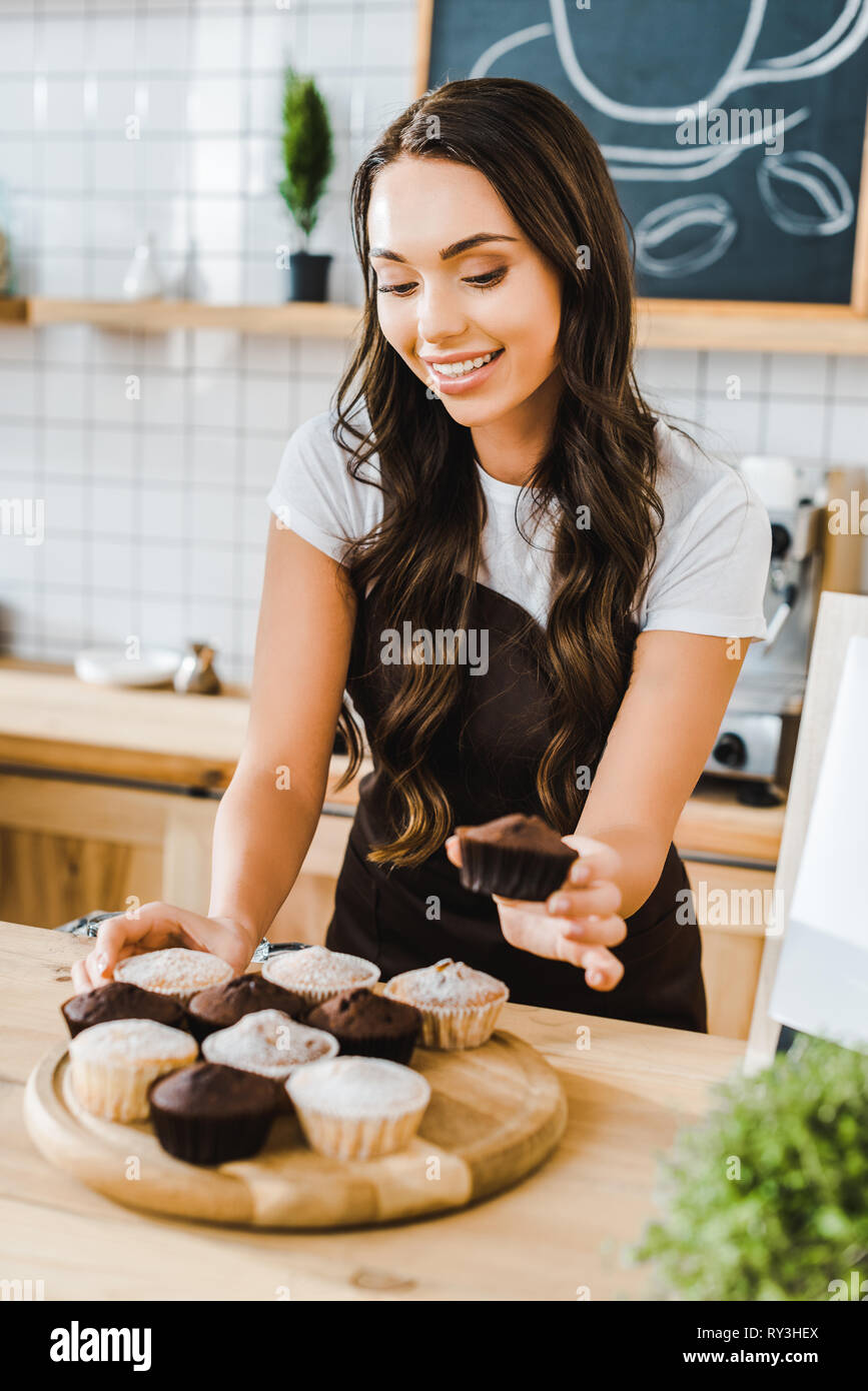 selective focus of attractive waitress standing behind bar counter and putting cupcakes on