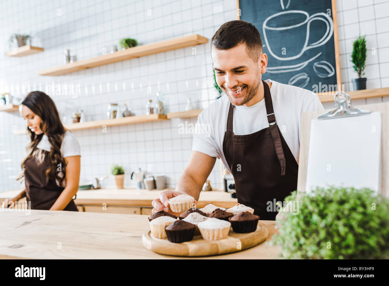 selective focus of waiter standing behind bar counter with cupcakes wile cashier working in coffee house Stock Photo