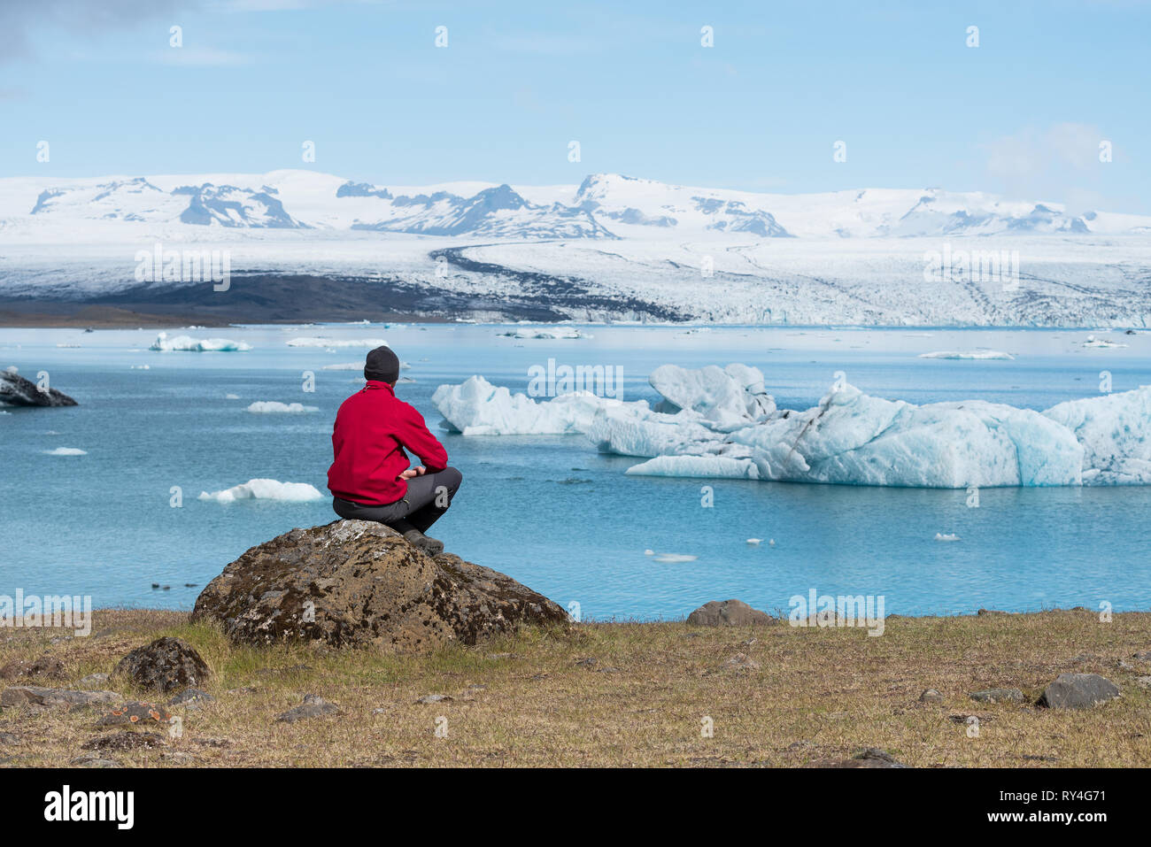 Man traveler in red jacket sits on the shore of a lake. Summer landscape with glacial lagoon, glacier and icebergs in the southeast of Iceland, Europe Stock Photo