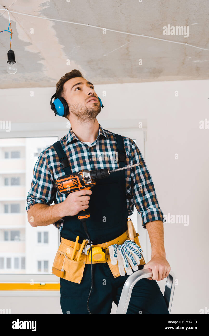 handsome handyman holding drill in hand and looking on ceiling Stock Photo