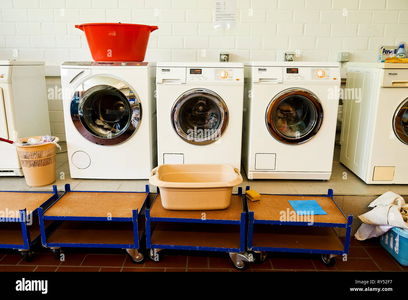 Waschmaschinen und ein Trockner stehen nebeneinader in einem Waschkeller. Auf dem Trockner steht ein roter Waeschekrob und im Vordergrund  eine Schues Stock Photo