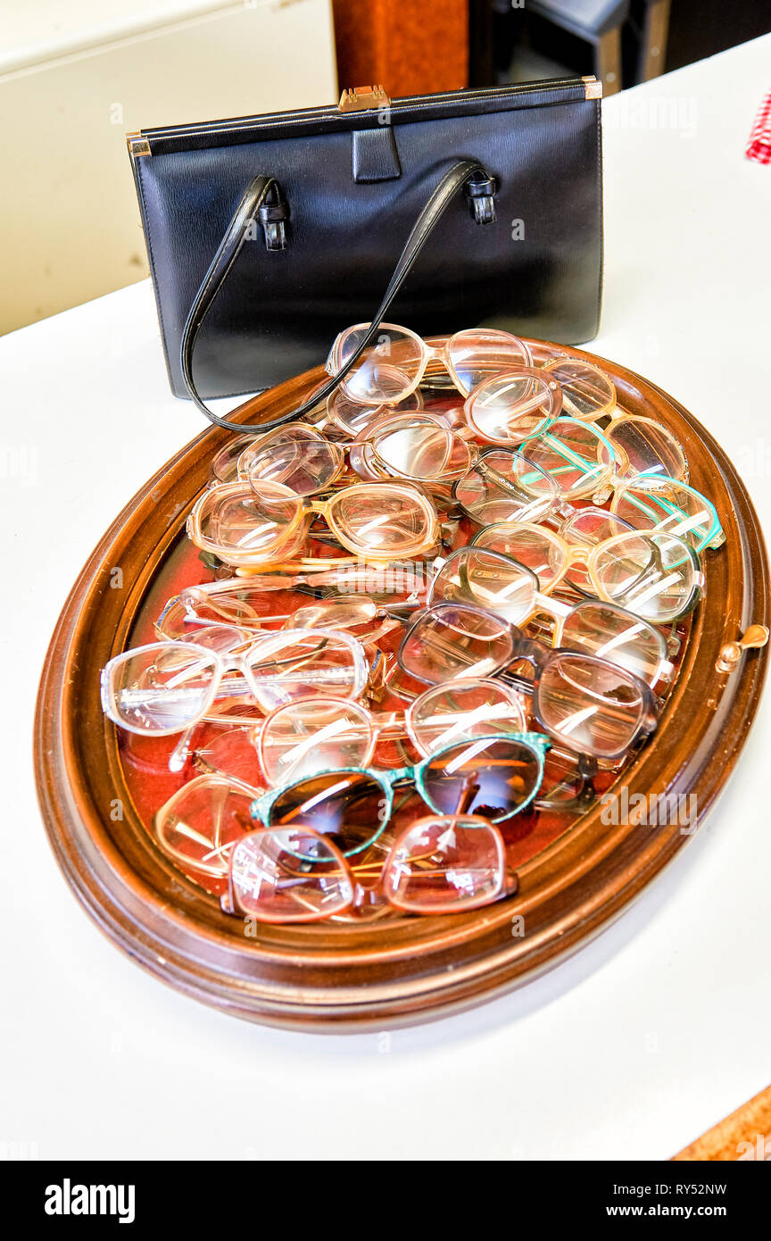Auf einem Bilderrahmen liegen viele verschiedene Brillengestelle in einem Gebrauchtwarenlgeschaeft. Dahinter steht eine schwarze Damenledertasche Stock Photo