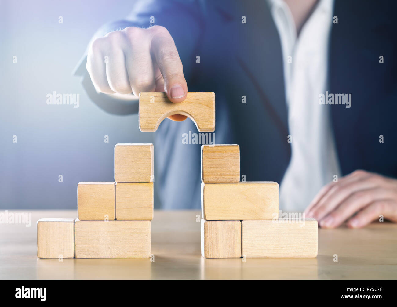 Business man bridging the gap between two towers or parties made from wooden blocks; conflict management or mediator concept, blue toned with ligth fl Stock Photo