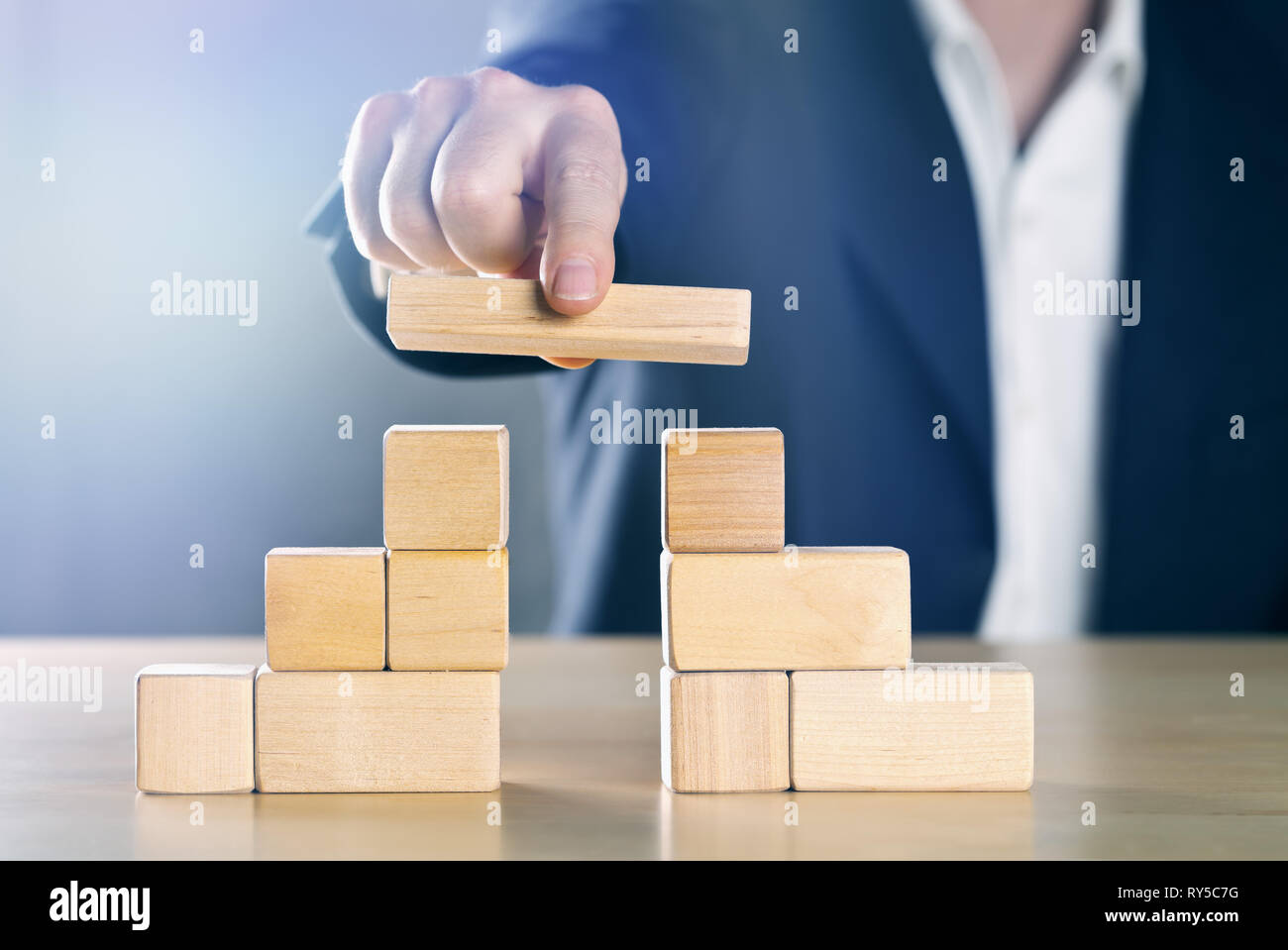 Business man bridging the gap between two towers or parties made from wooden blocks; conflict management or mediator concept, blue toned with ligth fl Stock Photo