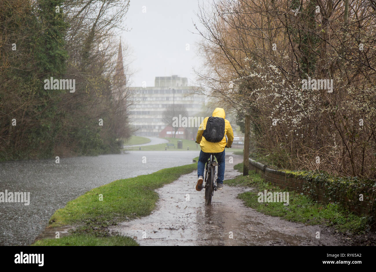 Kidderminster, UK. 12th March, 2019. UK weather: heavy rain throughout the UK leaves some commuters finding the going a little tough! An isolated male cyclist in yellow hi-vis jacket, seen here from behind, cycles along a canal towpath unable to avoid the muddy puddles forming as torrential rain persists. Credit Lee Hudson/Alamy Live News Stock Photo