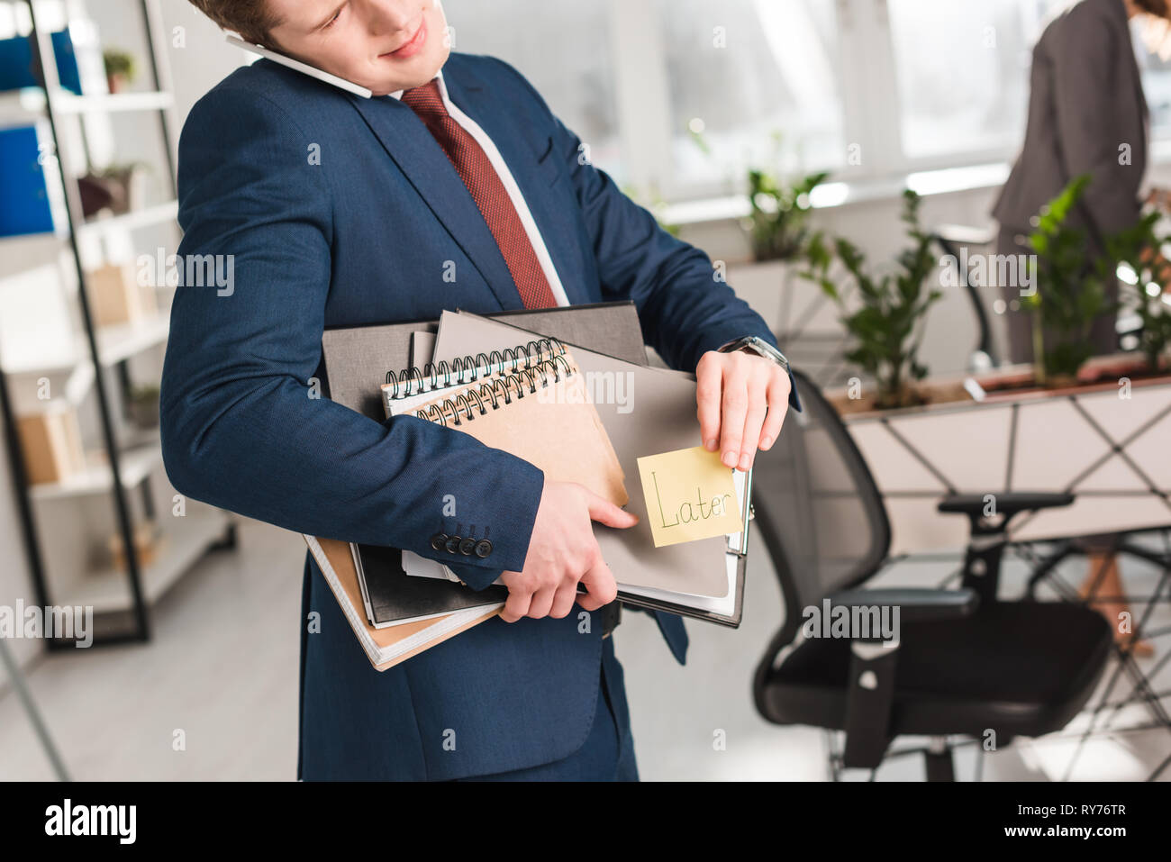 cropped view of businessman holding folders and sticky note with later lettering and talking on smartphone in office Stock Photo