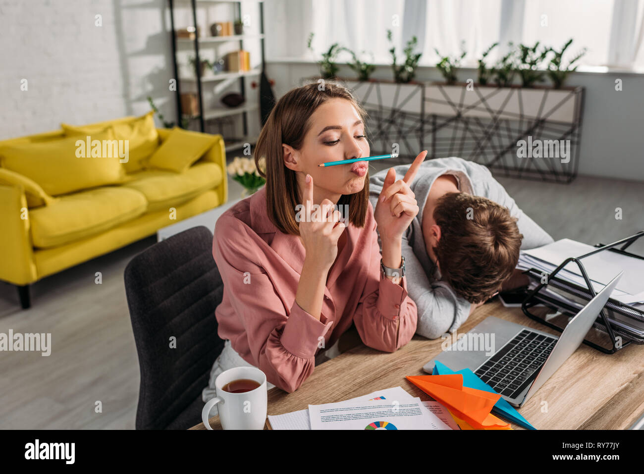 funny girlfriend sitting near boyfriend sleeping at desk near laptop and cup with drink Stock Photo