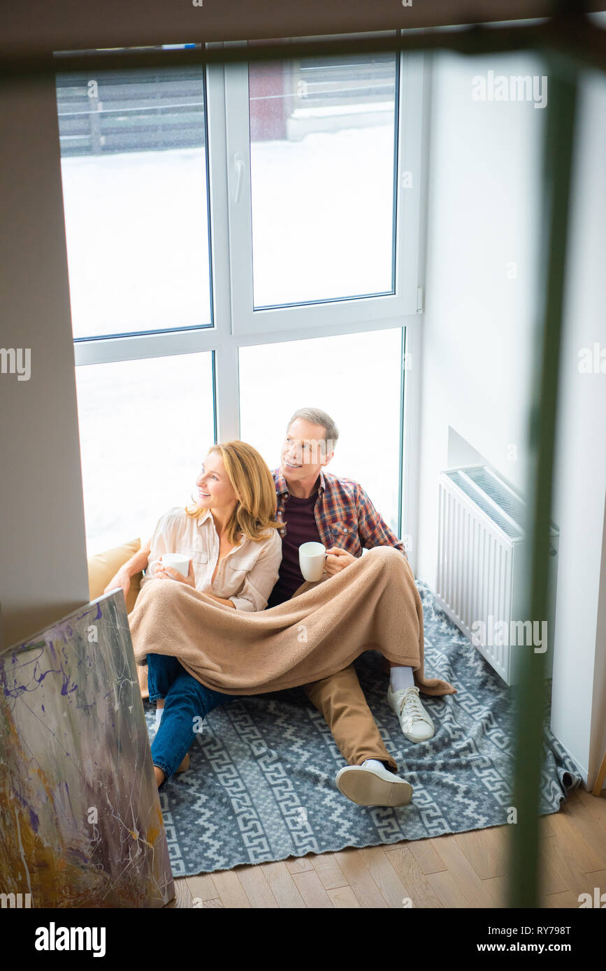 happy couple sitting on floor by large window and holding coffee cups ...