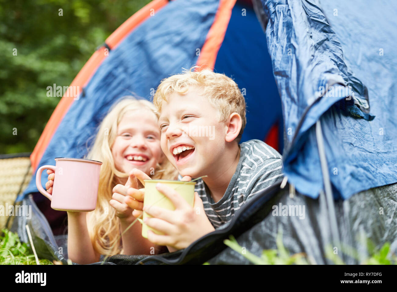 Two laughing kids with cups look out of their tent while camping Stock Photo