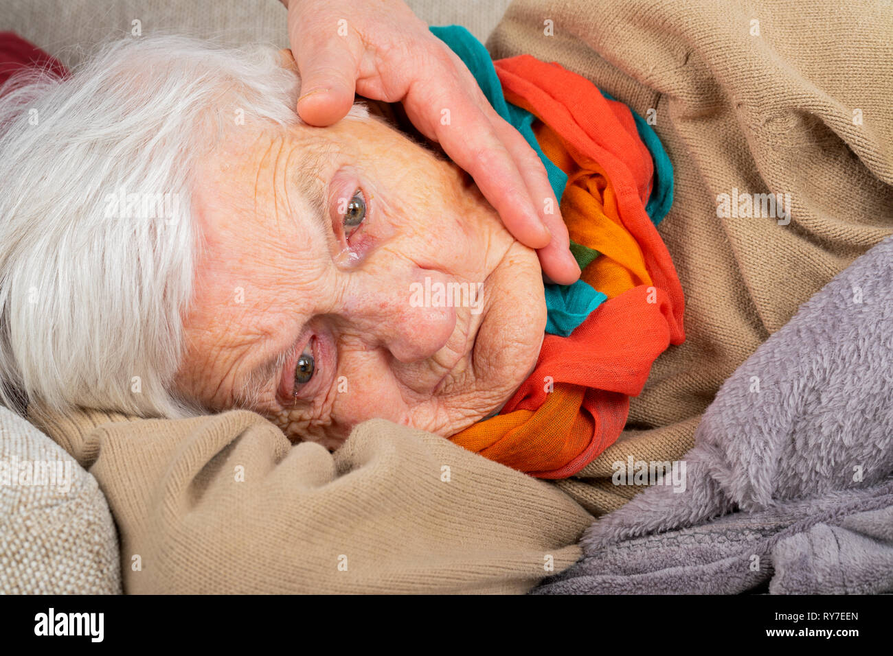 Close up portrait of a sad elderly wrinkled woman lying on the couch, wearing colorful scarf - caregiver's hand touching her face Stock Photo
