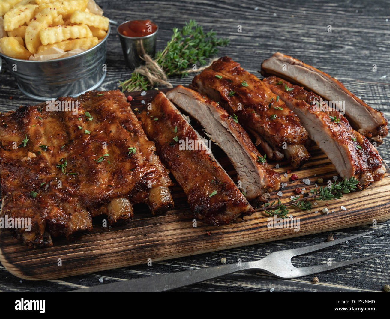 cutting grilled pork ribs with sauce on a board, french fries, spices, wooden background Stock Photo