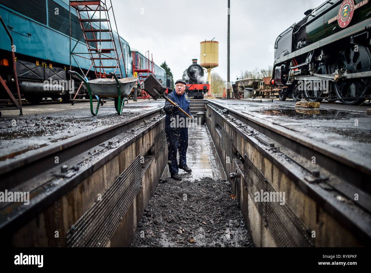 An engineer shovels ash from a locomotive firebox inside an inspection pit as locomotives are prepared for service at Gloucestershire Warwickshire Steam Railway station at Toddington, where a special heritage steam express train service is taking race goers to Cheltenham. Stock Photo