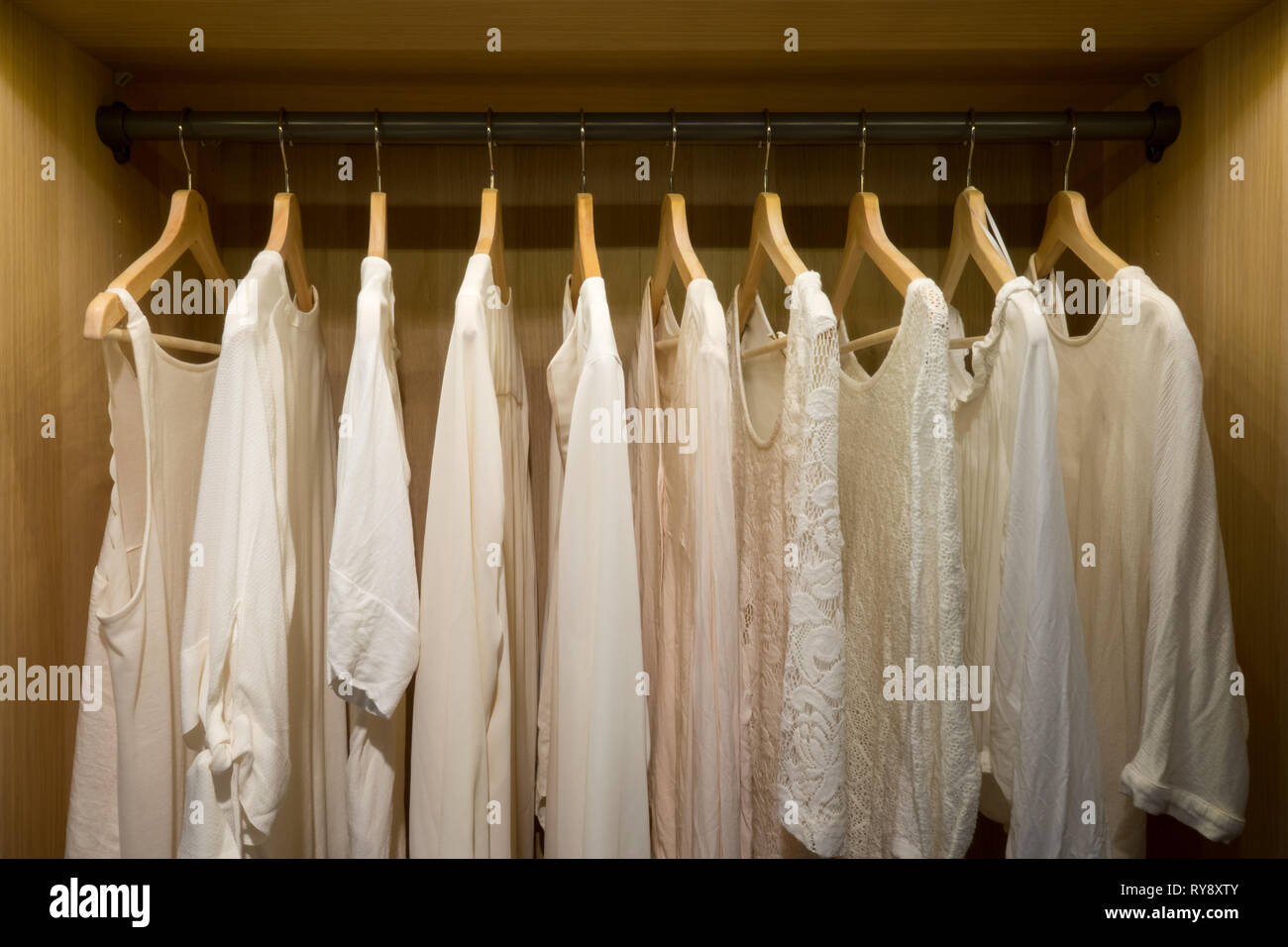 Picture taken straight into a wardrobe with lighting. White summer blouses and sweaters on hangers. Stock Photo
