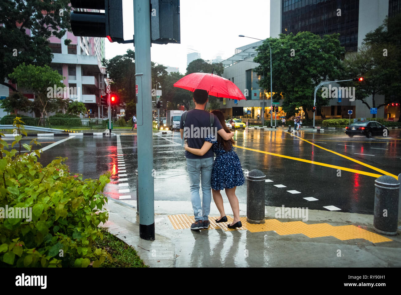 Young couple hugging under red umbrella at rainy crosswalk - Sim Lim Square - Singapore Stock Photo