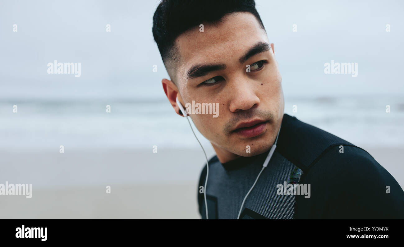 Close up shot of fit young man with earphone standing outside and looking away. Asian fitness model relaxing after physical workout session outdoors. Stock Photo