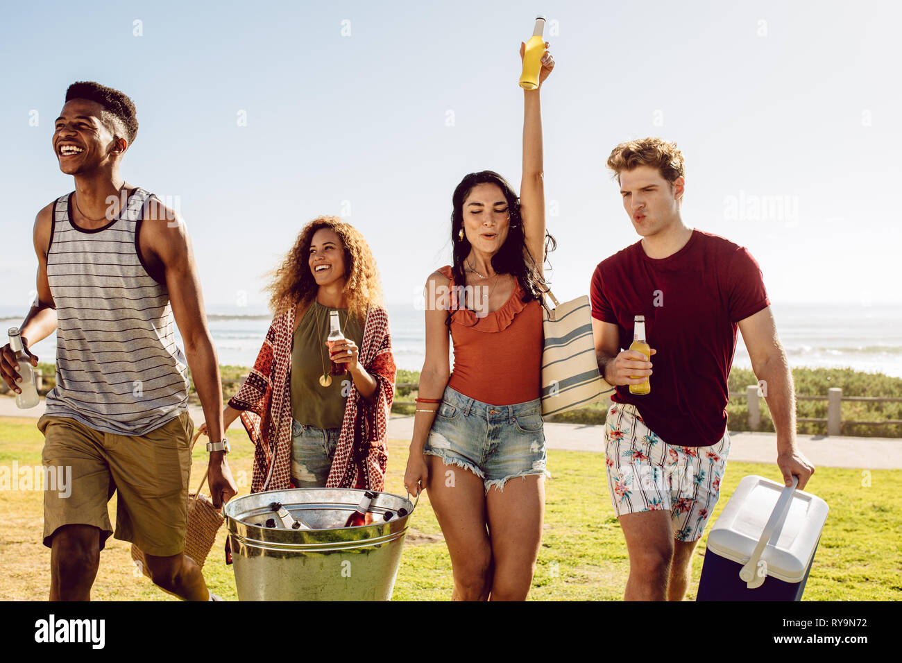 Friends carrying cooler and beverage tub for a picnic on beach. Multi-ethnic group of young people walking outdoors with drinks. Stock Photo
