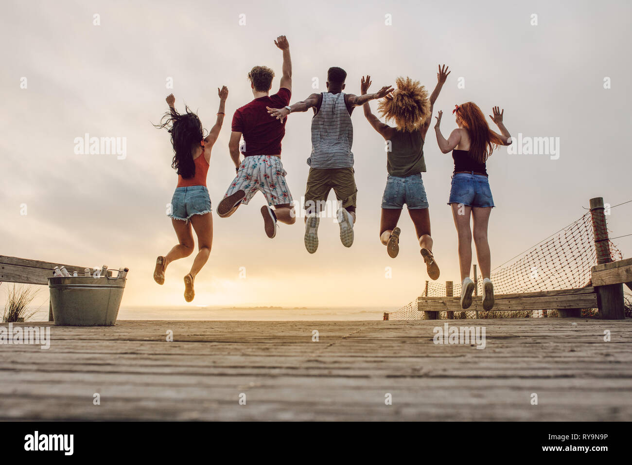 Rear view of young people jumping at the beach during sunset. Group of friends enjoying at the beach. Stock Photo
