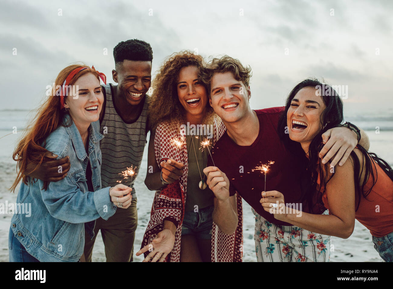 Multiracial friends enjoying at beach with sparklers. Group of young men and women having fun with fireworks at the sea shore. Stock Photo