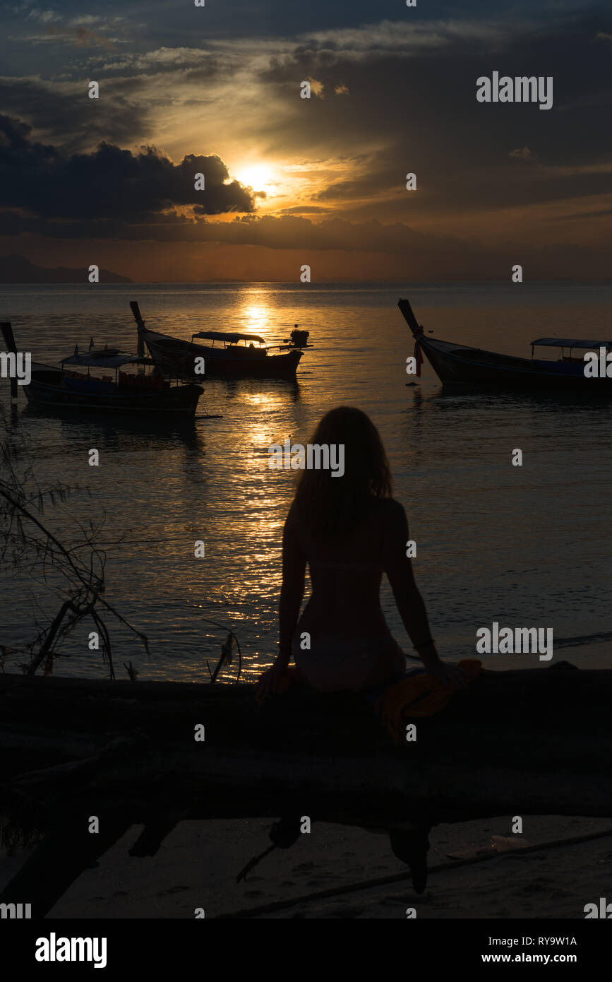 Silhouette of a girl sitting on a fallen tree on Sunrise beach against rising sun, Ko Lipe, Thailand Stock Photo
