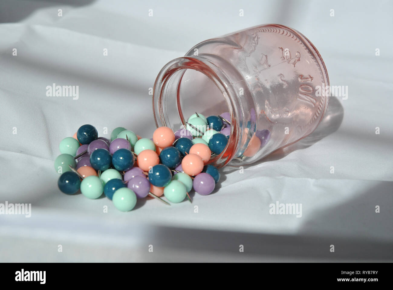 A jar of push pins on its side with pins spilling out, against a white