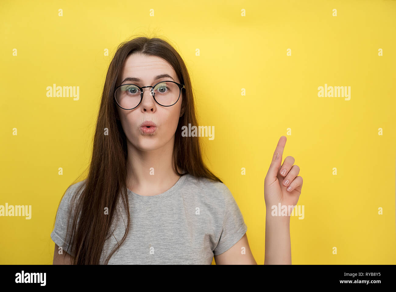 Surprised Caucasian young lady has bated breath, dressed in gray T-shirt, models against yellow background, demonstrates something at blank copy space Stock Photo