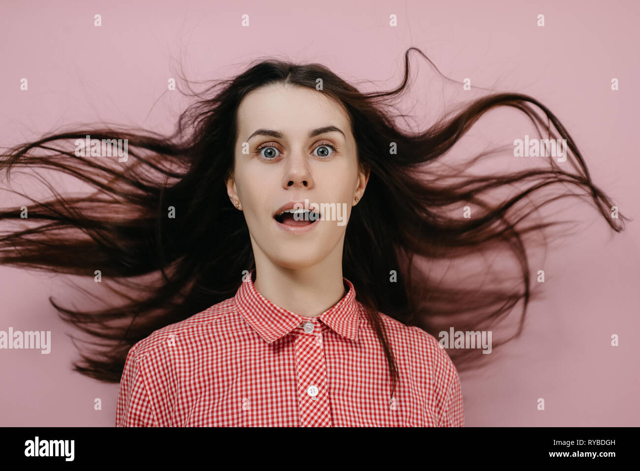 Shocked young girl with flying hair in panic looks with surprised expression, wears red shirt, opens mouth with stupefaction, isolated on pink wall. Stock Photo
