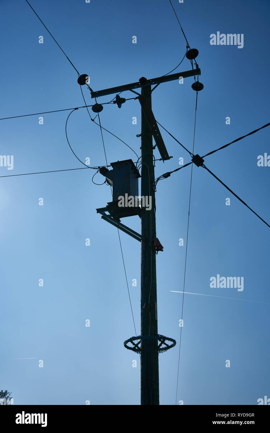Silhouette of a wooden electrical power distribution post and overhead cables against a blue sky. Stock Photo