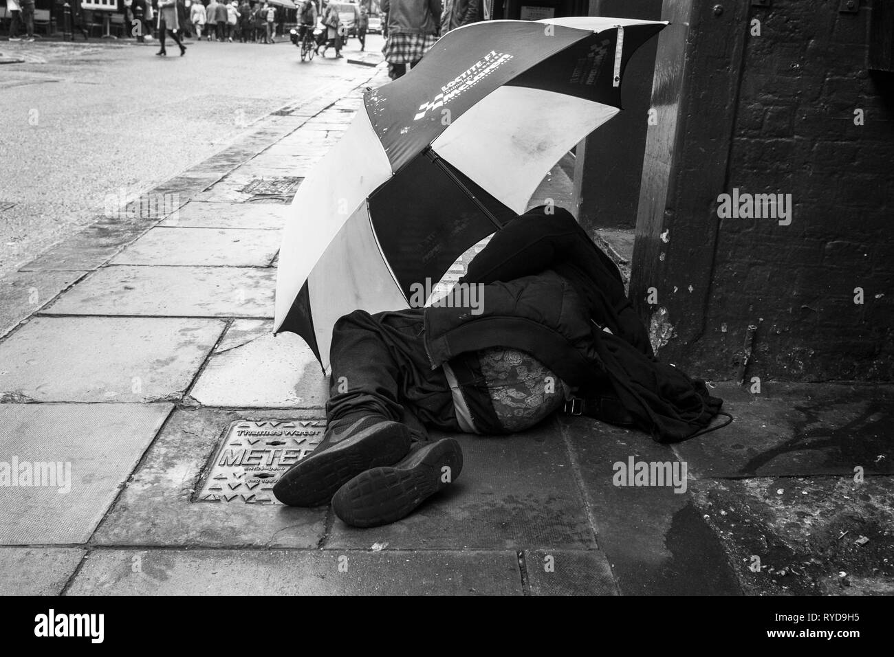 London black and white street photography: Homeless man using large umbrella for shelter on central London street. UK Stock Photo