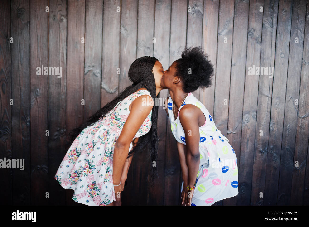 Two black african girlfriends at summer dresses posed against dark wooden b...