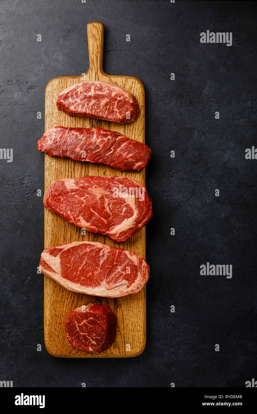 Variety of Raw Black Angus Prime meat steaks Top Blade, Denver, Rib Eye, Striploin, Tenderloin fillet Mignon on wooden board on dark background Stock Photo