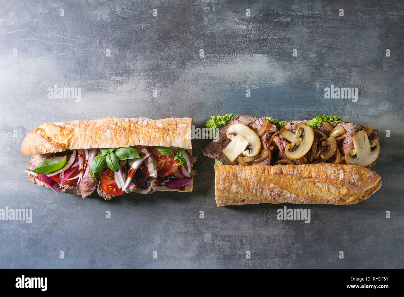 Two Beef baguette sandwiches with champignon mushrooms, green salad, fried onion, tomatoes over blue texture background. Flat lay, space Stock Photo