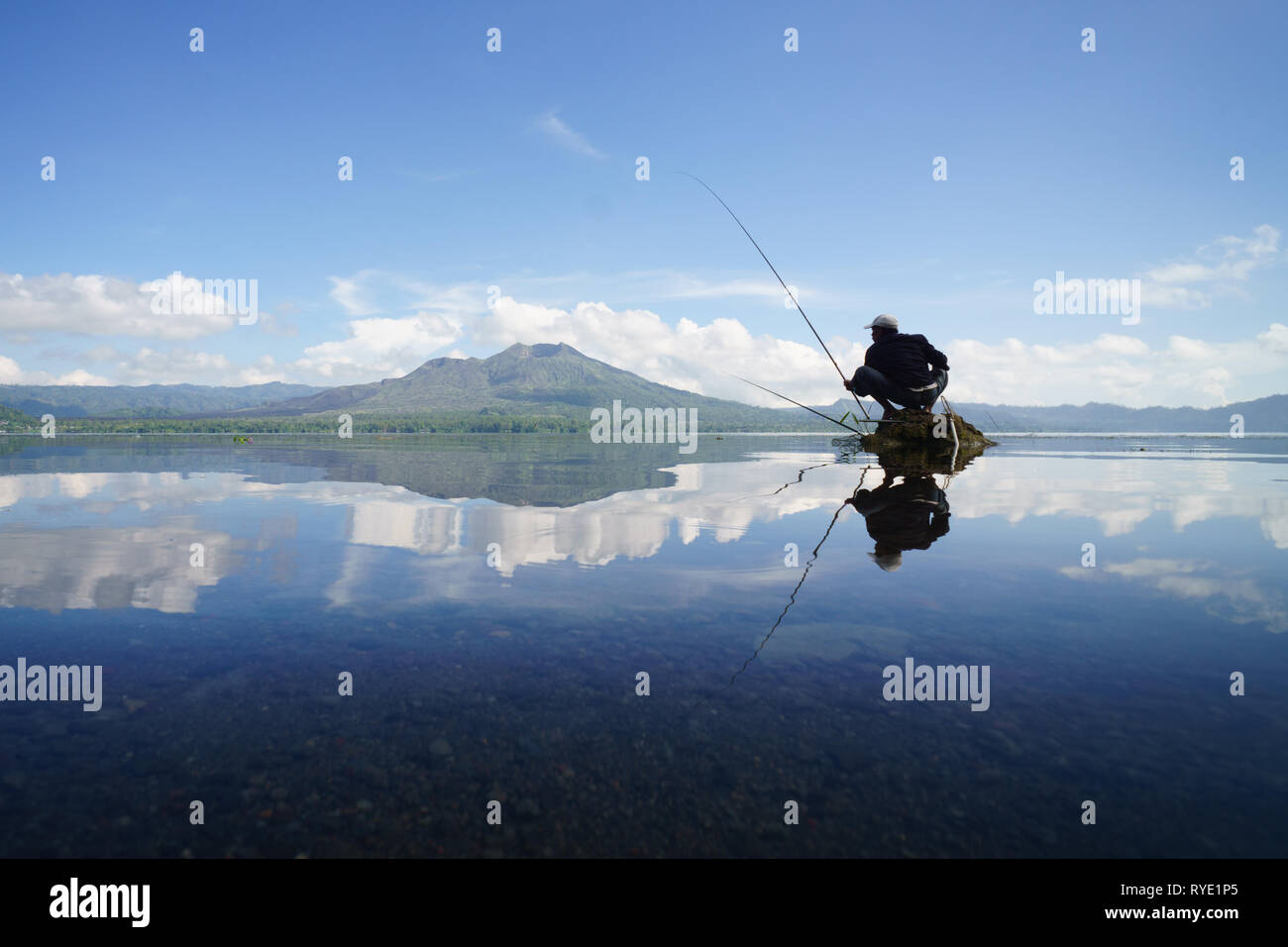 Symmetry reflection of Asian villager man fishing on top of rock in the middle of crystal clear lake with scenic panorama. Stock Photo