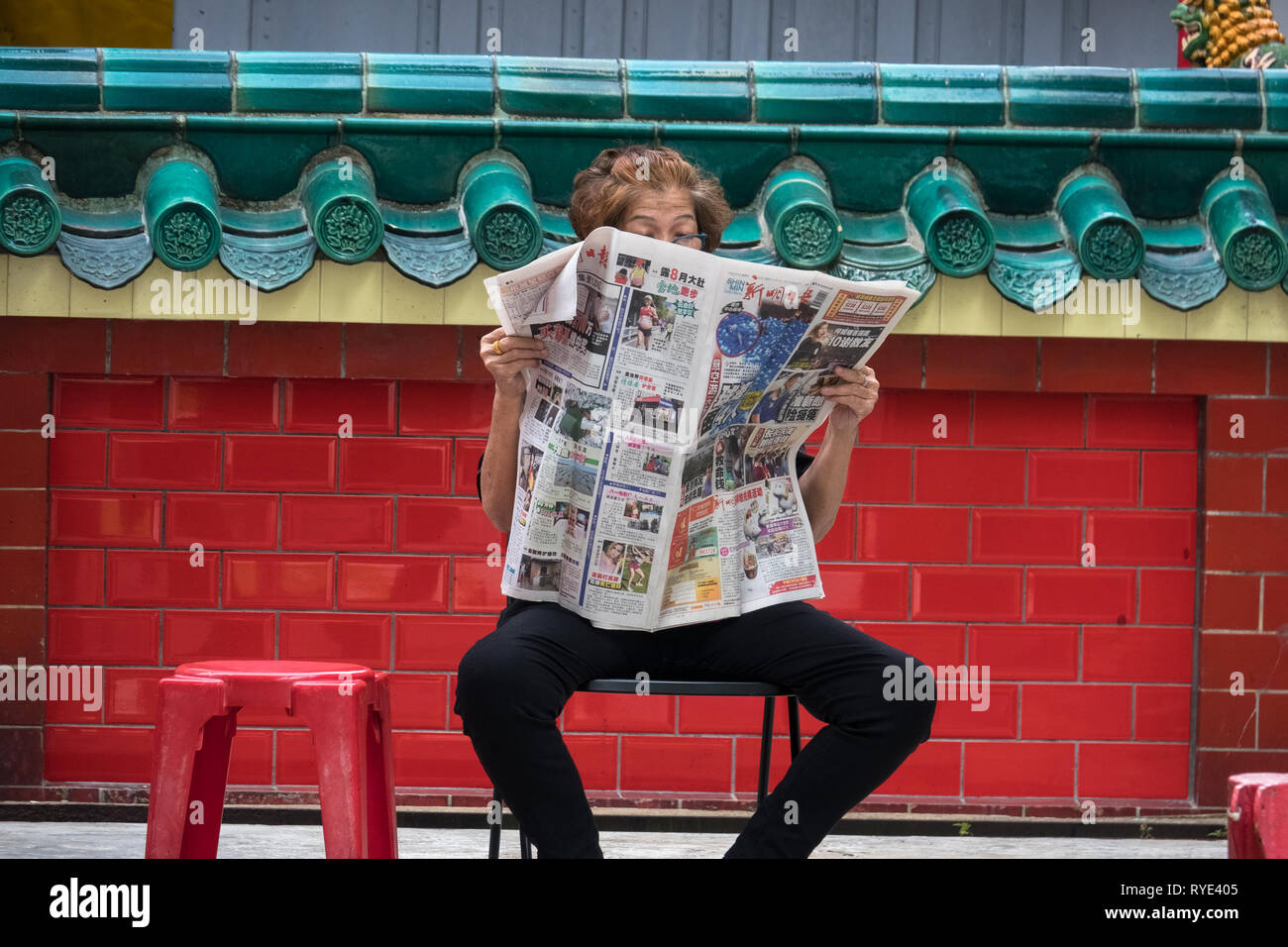Old Singaporean woman reading newspaper on chair at Chinese temple - Singapore Stock Photo