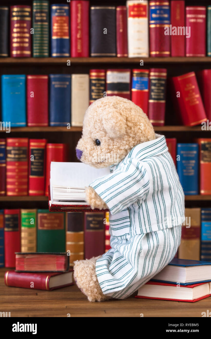 Cute teddy bear in nostalgic striped pajamas sitting on stack of books in front of bookshelf, reading at open book in his hands Stock Photo
