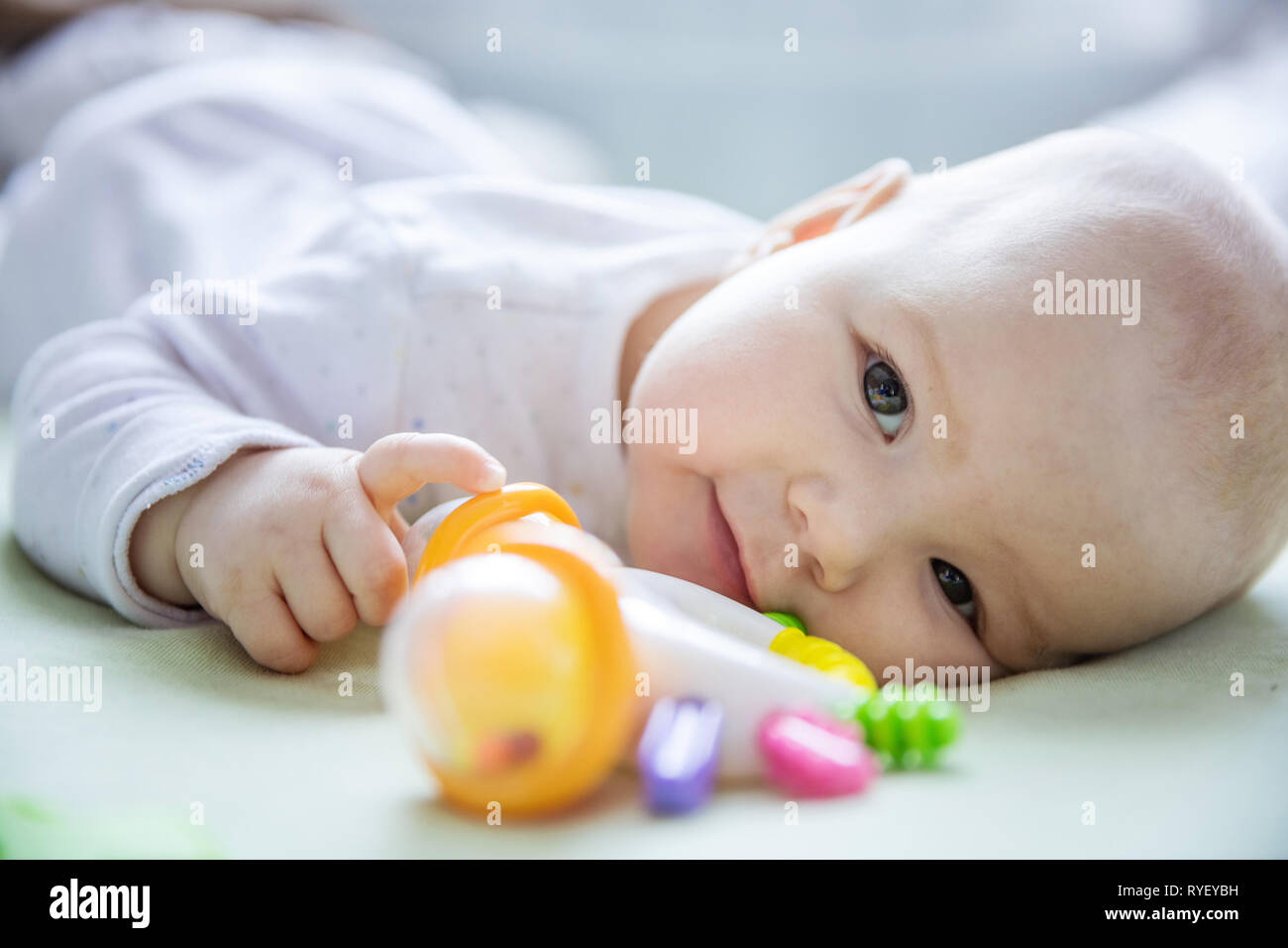 Cute baby girl playing with rattle while lying down on bed at home, closeup view Stock Photo