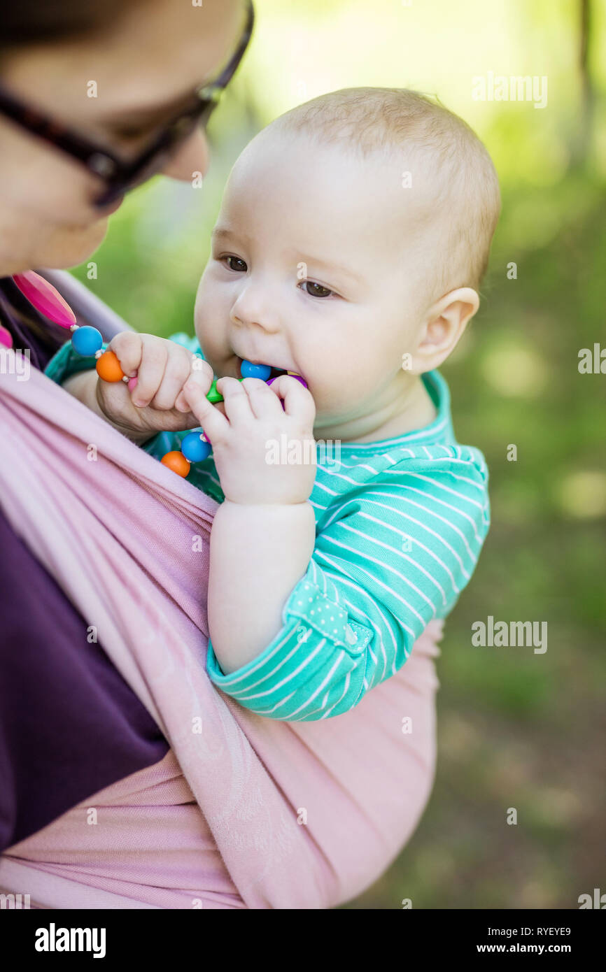 Young woman carrying her baby daughter in woven wrap outdoors in spring park. Baby girl chewing teething beads, mother looking at her with affection. Stock Photo