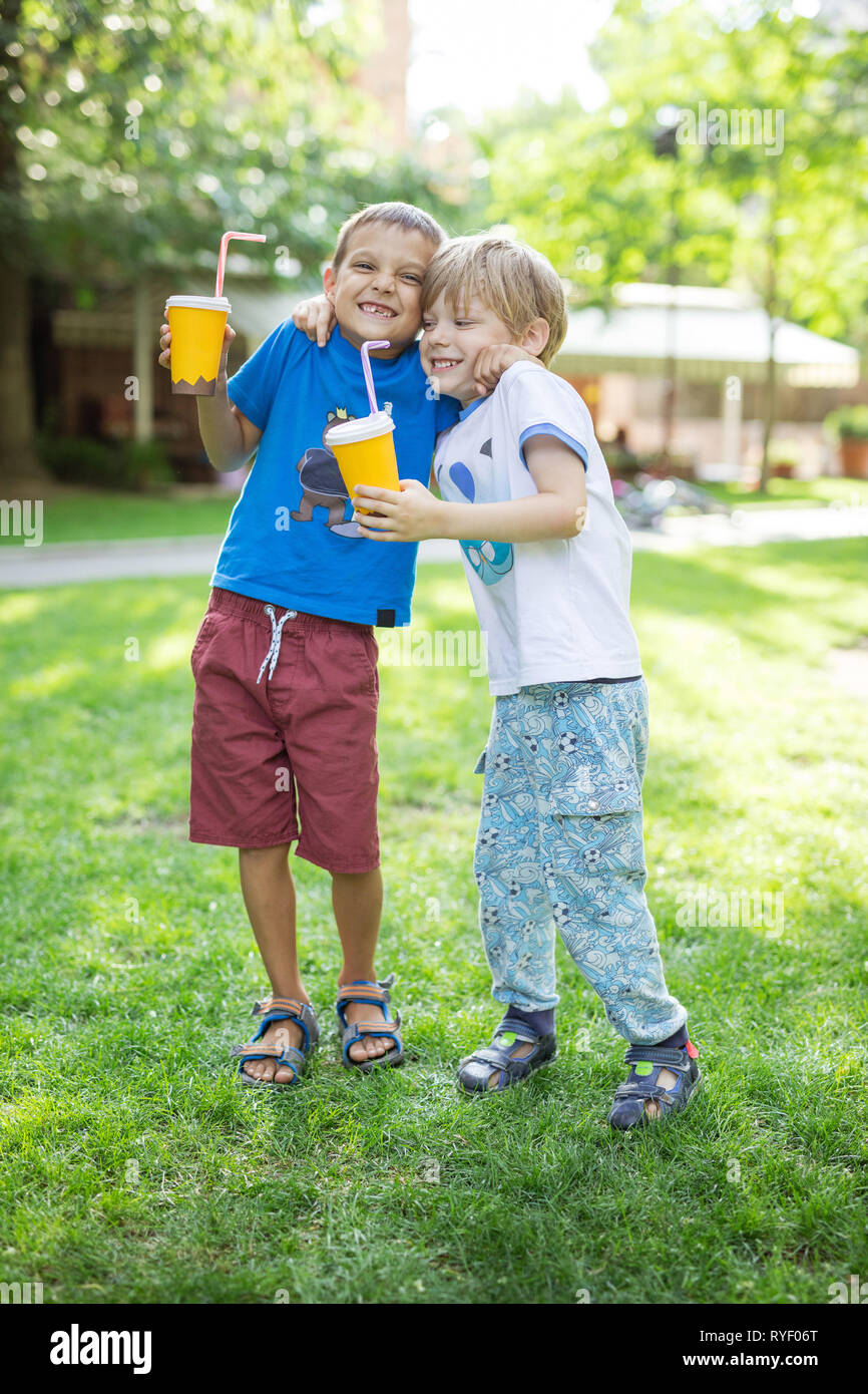 Two boys hugging one another or fighting jokingly while drinking cocoa from paper cups in park Stock Photo