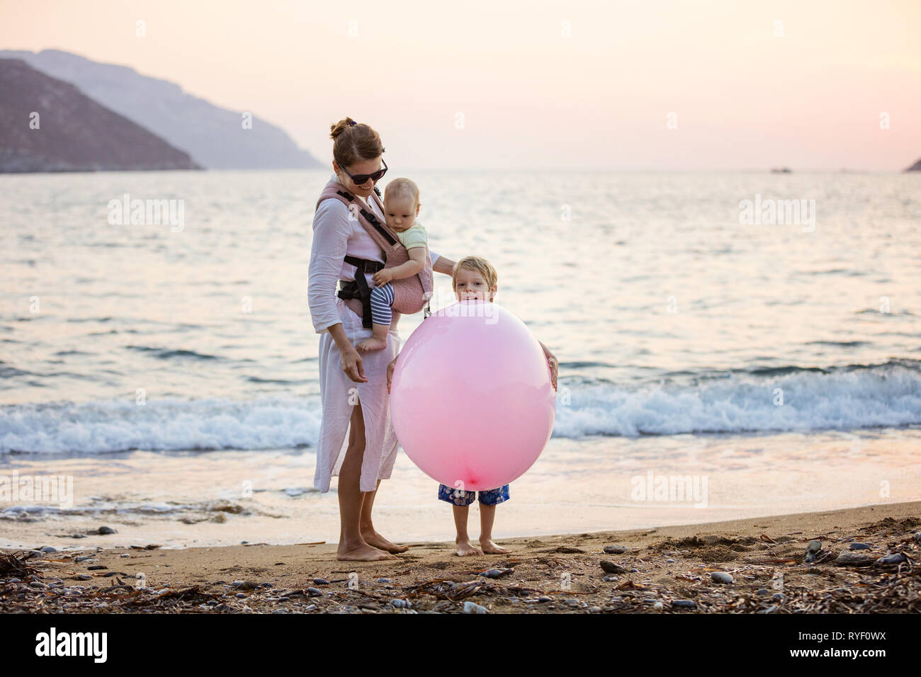 Young Caucasian woman with baby daughter in buckle carrier and preschool son playing with huge pink balloon on beach at sunset Stock Photo