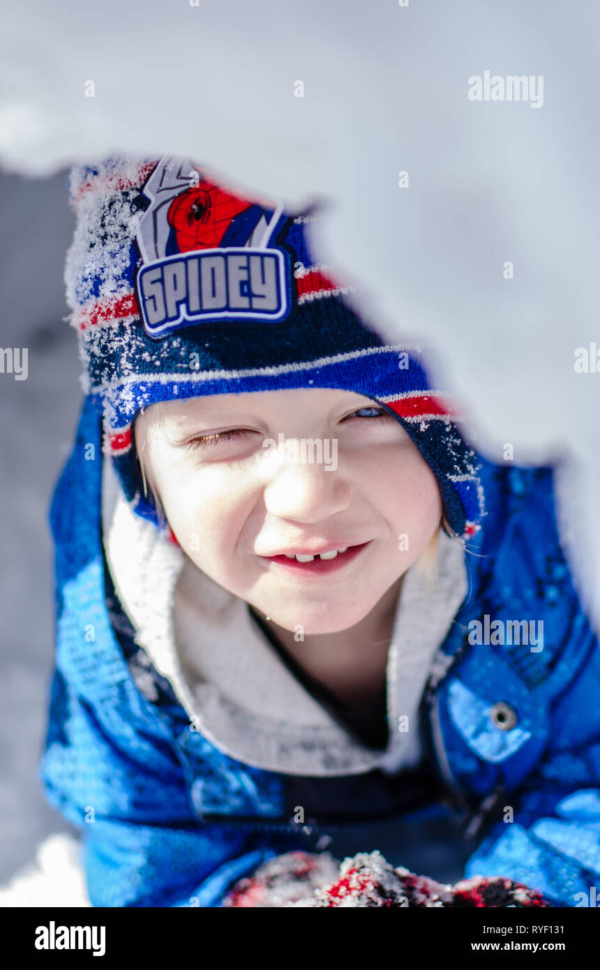 Cute toddler smiling hiding in the snow during a sunny winter day in Minnesota. The kid is wearing a blue jacket and a funny hat. Stock Photo