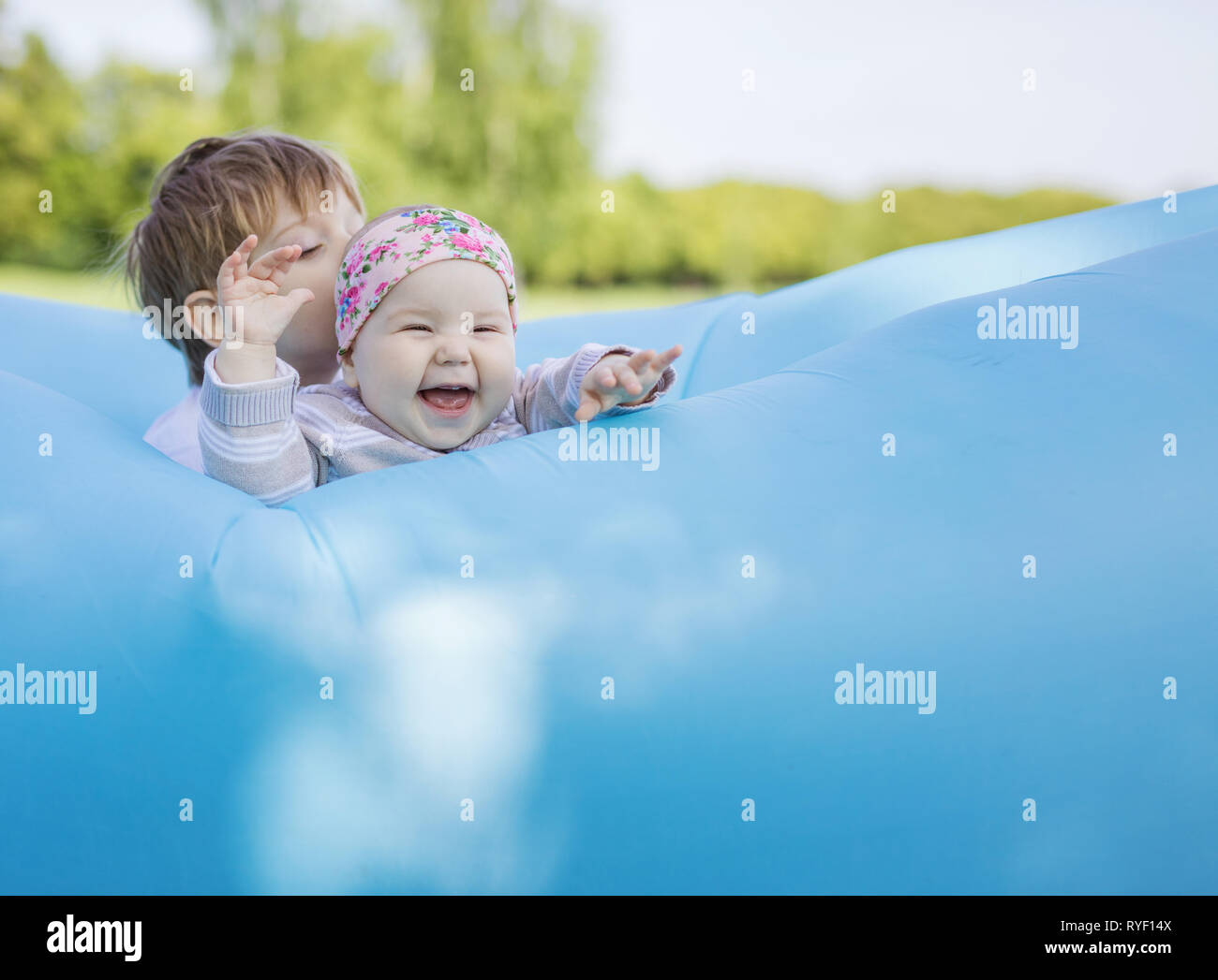 Siblings playing on inflatable sofa outdoors. Baby girl and older brother having fun together in park. Stock Photo