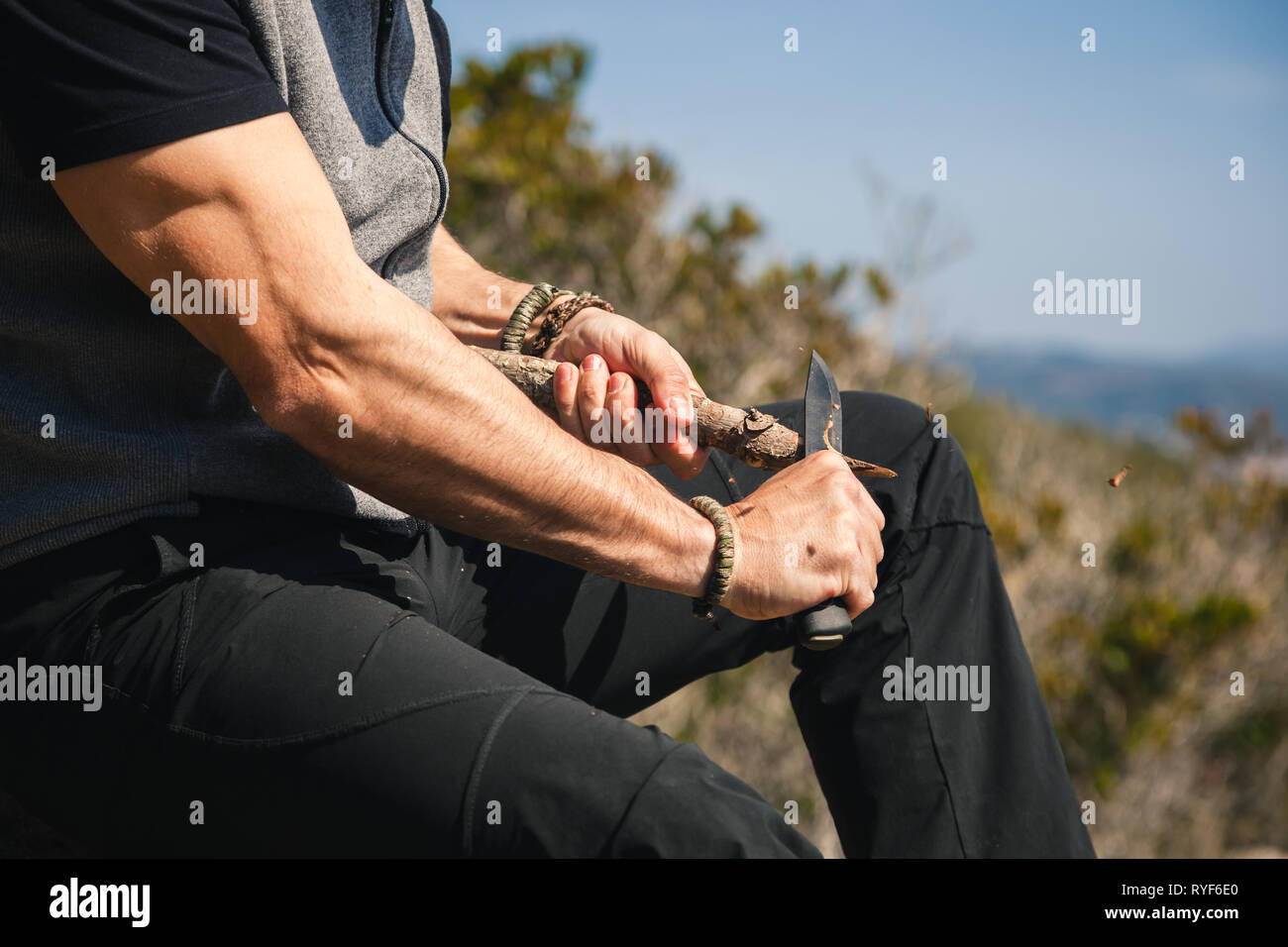 Man sitting on a rock in the nature with great view carving a stock of a tree with a knife, closeup Stock Photo